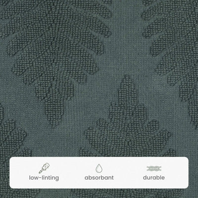 Close-up of a jade bath rug textured fabric with a subtle tonal leaf pattern. A white label at the bottom lists features: low-linting, absorbent, and durable.