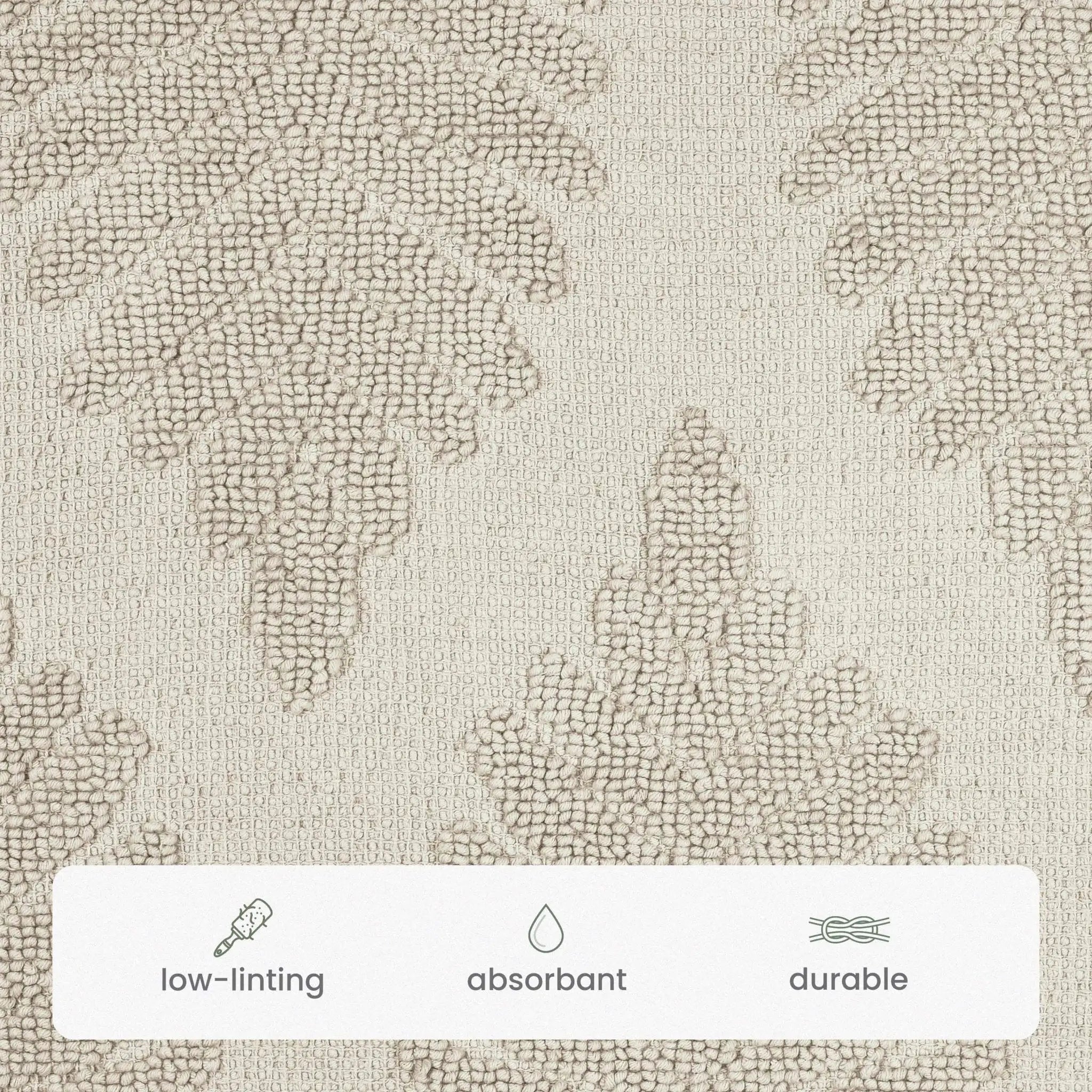 Close-up of a textured bath rug fabric swatch in sand color with a subtle leaf-like pattern, plus a white label at the bottom with icons reading low-linting, absorbent, and durable.
