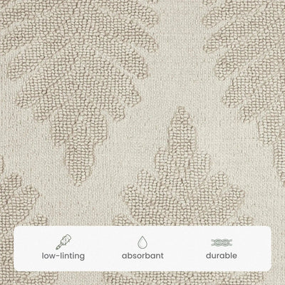 Close-up of a textured bath rug fabric swatch in sand color with a subtle leaf-like pattern, plus a white label at the bottom with icons reading low-linting, absorbent, and durable.