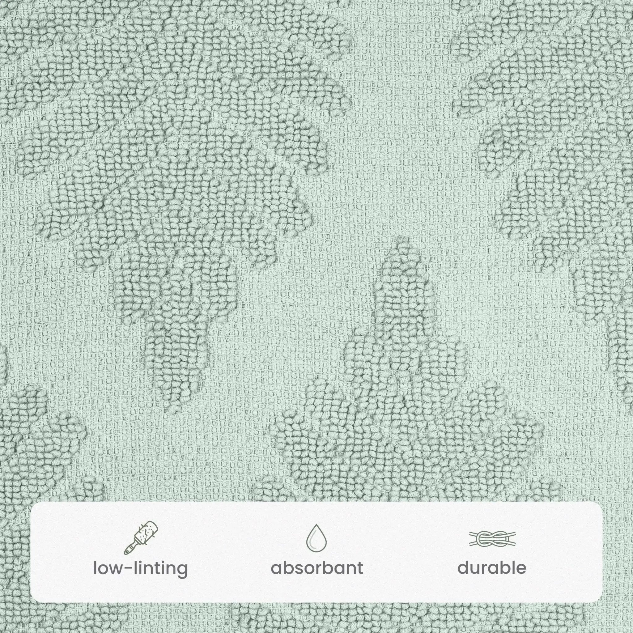 A close-up of a sage color bath rug with a subtle tonal damask pattern. A white banner at the bottom features icons and text highlighting key product features: 'low-linting', 'absorbant', and 'durable'.