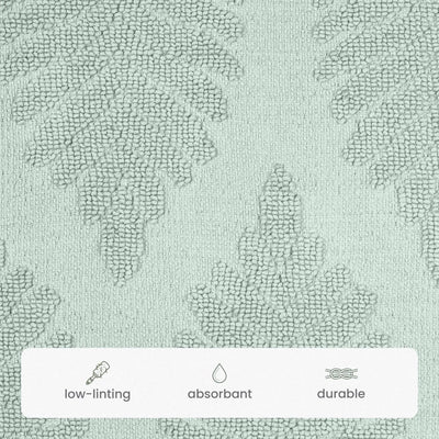 A close-up of a sage color bath rug with a subtle tonal damask pattern. A white banner at the bottom features icons and text highlighting key product features: 'low-linting', 'absorbant', and 'durable'.