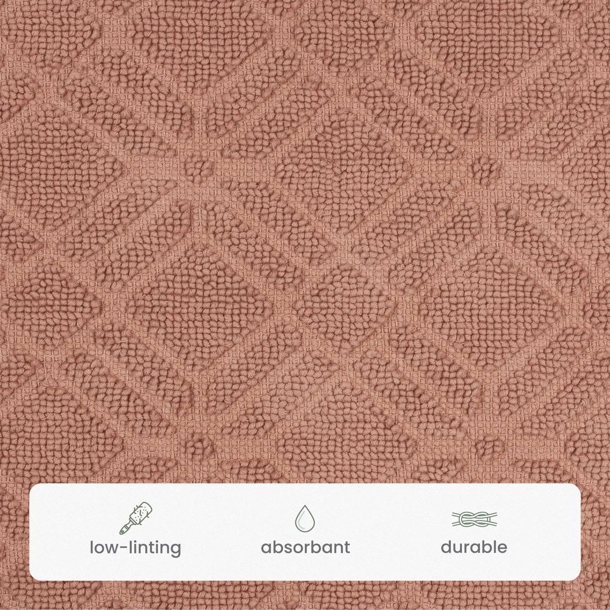 Close-up of a textured fabric surface in a clay colored bath rug, with a white banner at the bottom containing small icons and the words low-linting, absorbant, and durable.