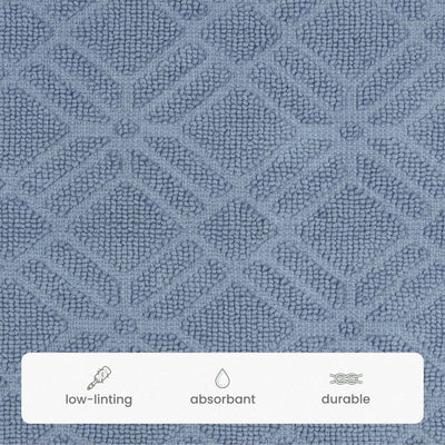 A close-up of a textured bath rug fabric swatch in a blue color, featuring a repeating geometric diamond pattern. A white banner at the bottom lists product features: 'low-linting', 'absorbant', and 'durable' with simple line icons.