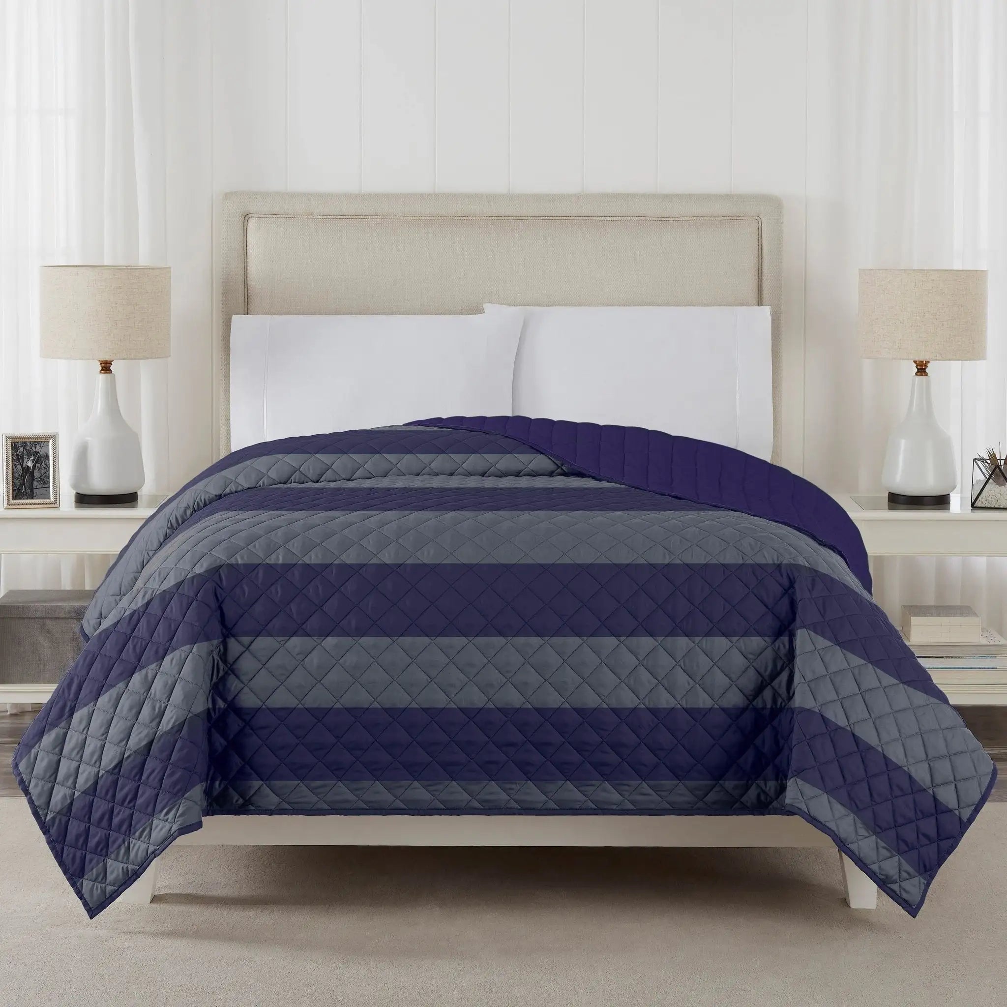 A striped quilted coverlet in shades of navy blue, medium blue, and gray, styled on a bed with a beige upholstered headboard, white pillows.