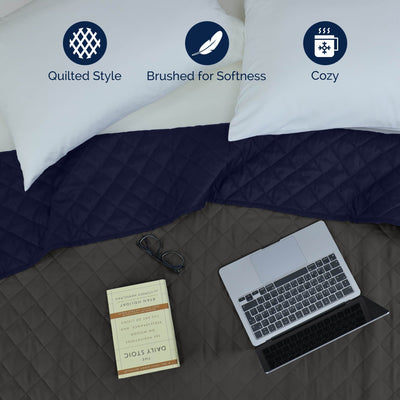With laptop, book, and glasses on a Graphite/navy coverlet, featuring icons for quilted style, coziness, and gentle on skin.