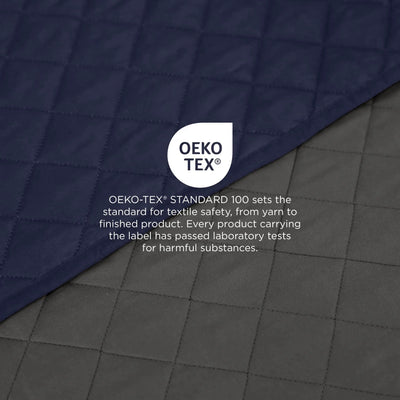 A close-up of quilted coverlet fabric in two tones: navy blue on top and dark gray below, with a centered OEKO-TEX logo and text explaining textile safety standards.