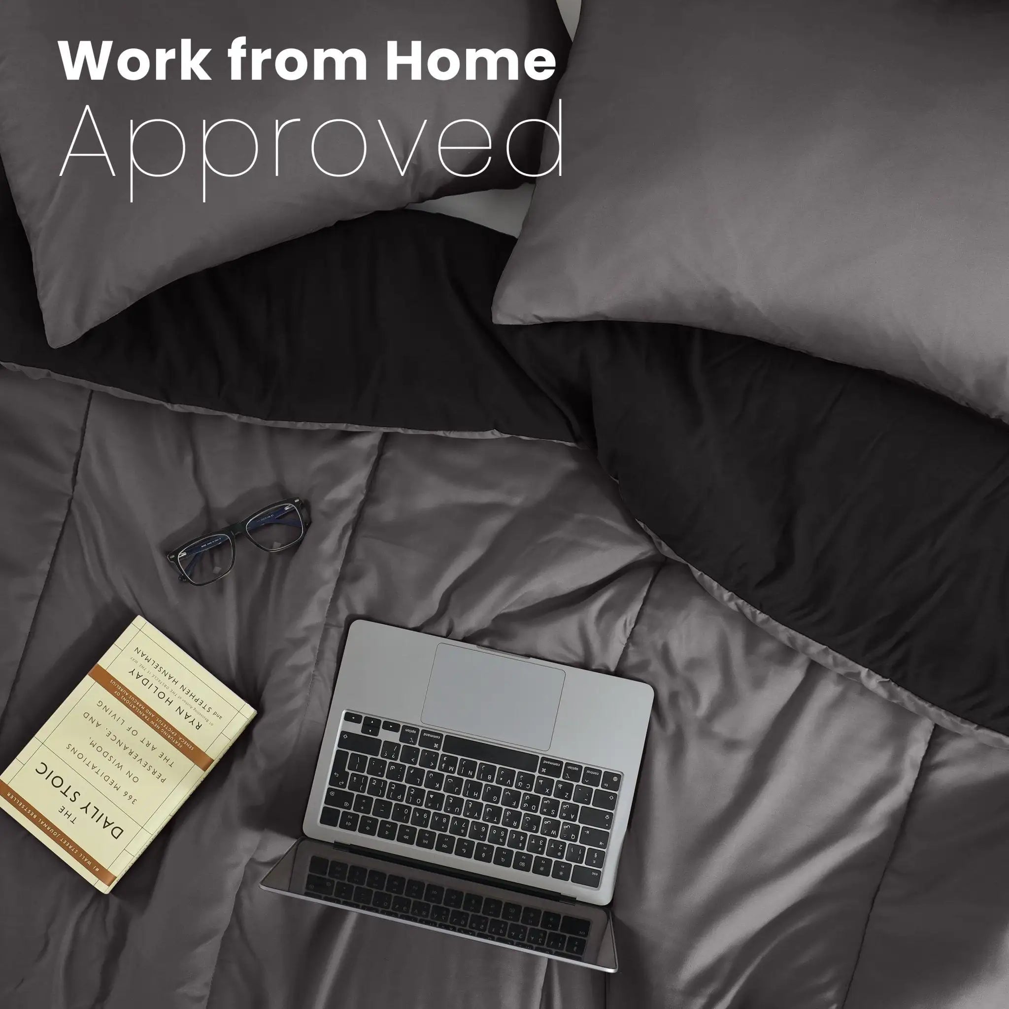 A top-down view of a graphite comforter and ebony reversible side with a laptop, glasses, and a book on the sheets, under the text 'Work from Home Approved'.