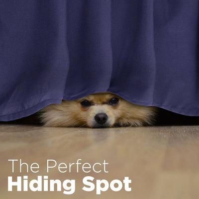 A small fluffy dog peeks out from under a navy bed skirt on a light wood floor, with the text 'The Perfect Hiding Spot' overlaid at the bottom.