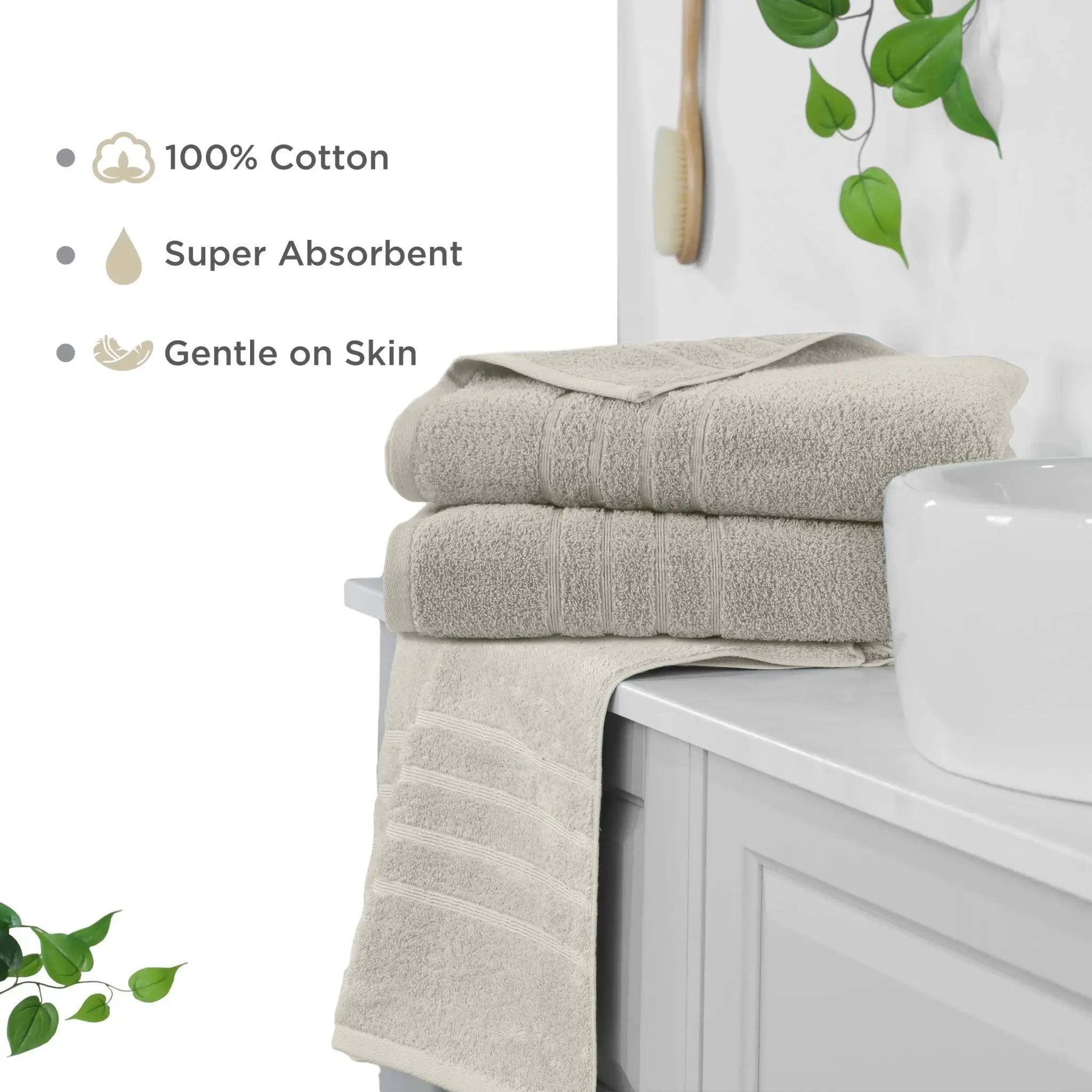 Three folded sand-colored cotton bath sheets stacked on a white bathroom vanity. The scene includes marketing text reading '100% Cotton', 'Super Absorbent', and 'Gentle on Skin', along with decorative green leaves and a sink.