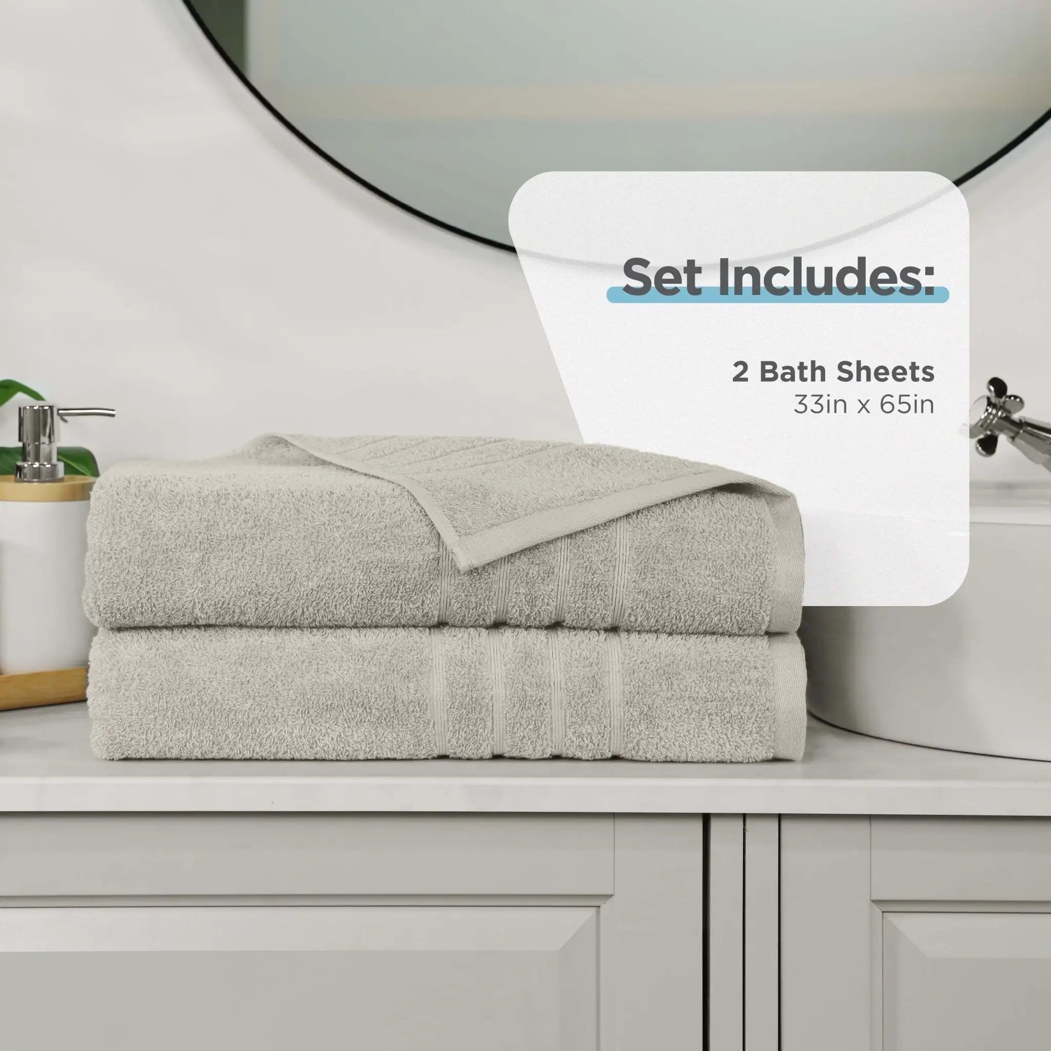 Two folded sand color bath sheets stacked on a bathroom counter, with a white overlay that reads 'Set Includes: 2 Bath Sheets 33in x 65in'. A sink, mirror, soap dispenser, and cabinet are visible in the background.