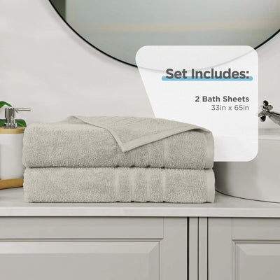 Two folded sand color bath sheets stacked on a bathroom counter, with a white overlay that reads 'Set Includes: 2 Bath Sheets 33in x 65in'. A sink, mirror, soap dispenser, and cabinet are visible in the background.