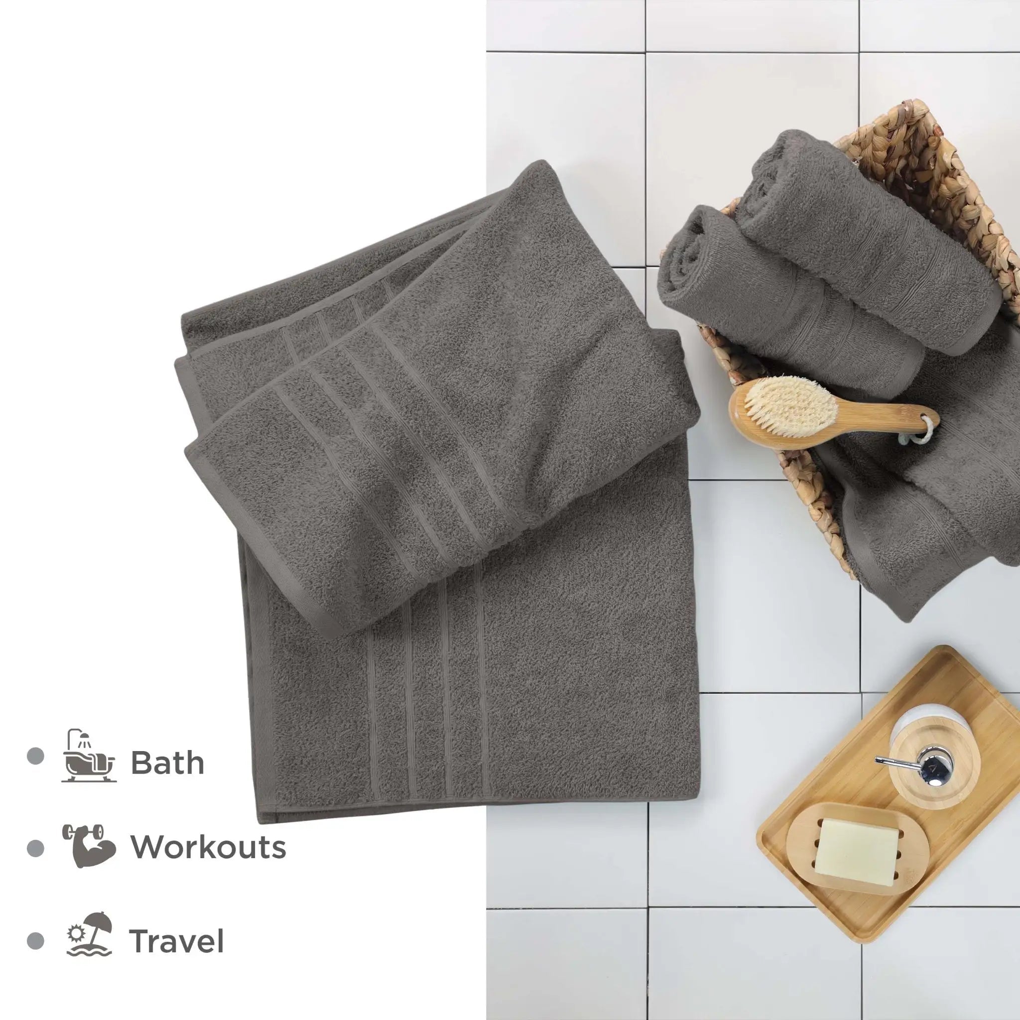 A set of dark gray cotton bath towels, including a large, folded towel and rolled towels in a woven basket, styled on a white tiled floor with a wooden brush. Text on the left lists uses: Bath, Workouts, and Travel.