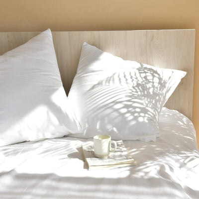 White pillows on a bed with a cup and book, bathed in sunlight.