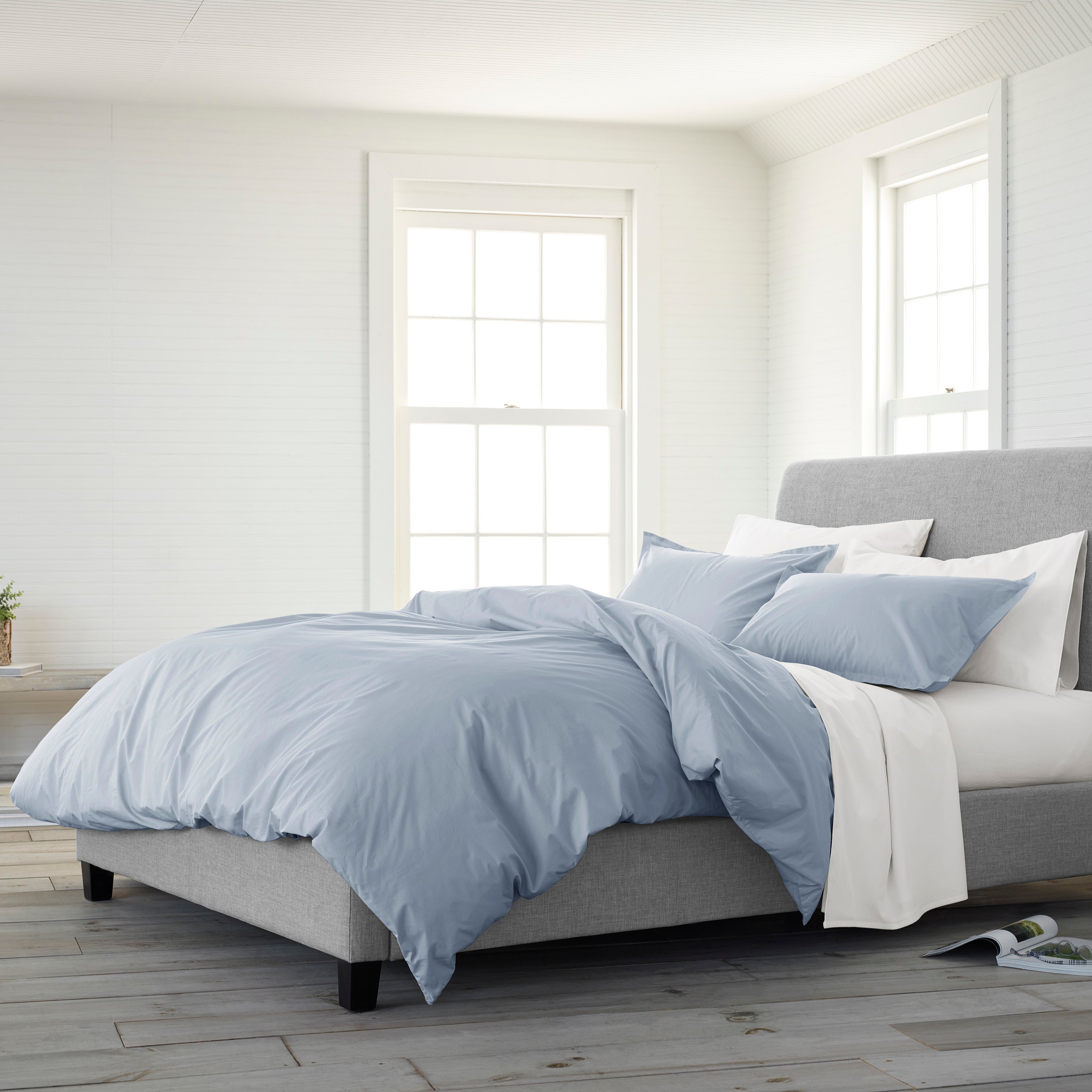 Light blue duvet set on a gray upholstered bed with white pillows, in a bright room with large windows.