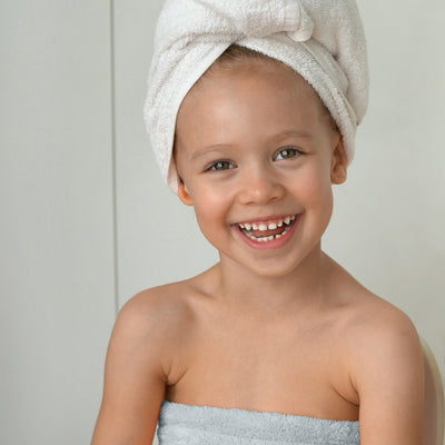 Child smiling while wearing a towel wrap and a towel on their head, seated against a light background.