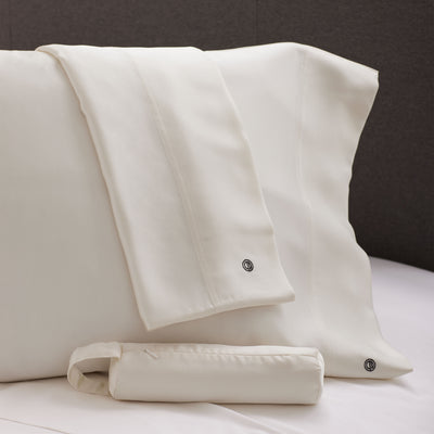 Two satin pillowcases in a light cream color with a small logo, placed on a bed.