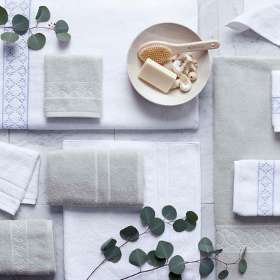 A collection of towels in white and light gray, arranged with a wooden bowl containing soap and a brush, and greenery.