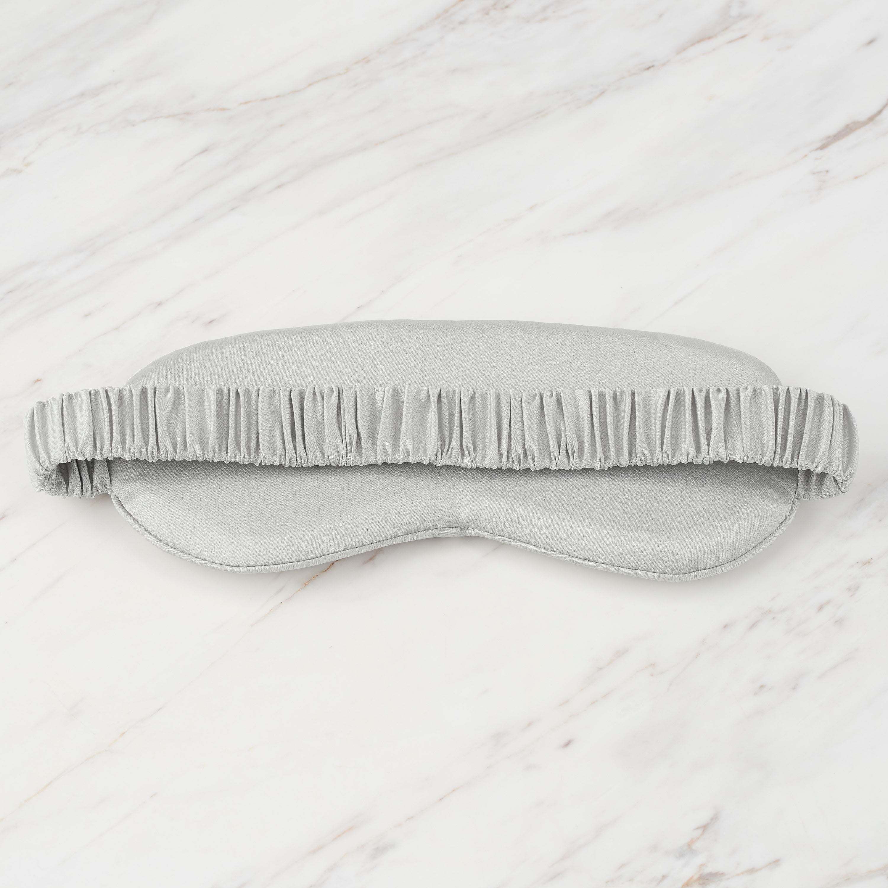 Gray satin eye mask with a ruffled elastic band, resting on a marble surface.