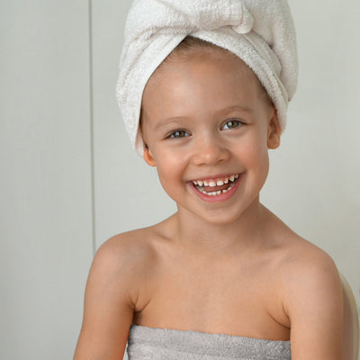 Child smiling while wearing a towel wrap and a towel on their head, seated against a light background.