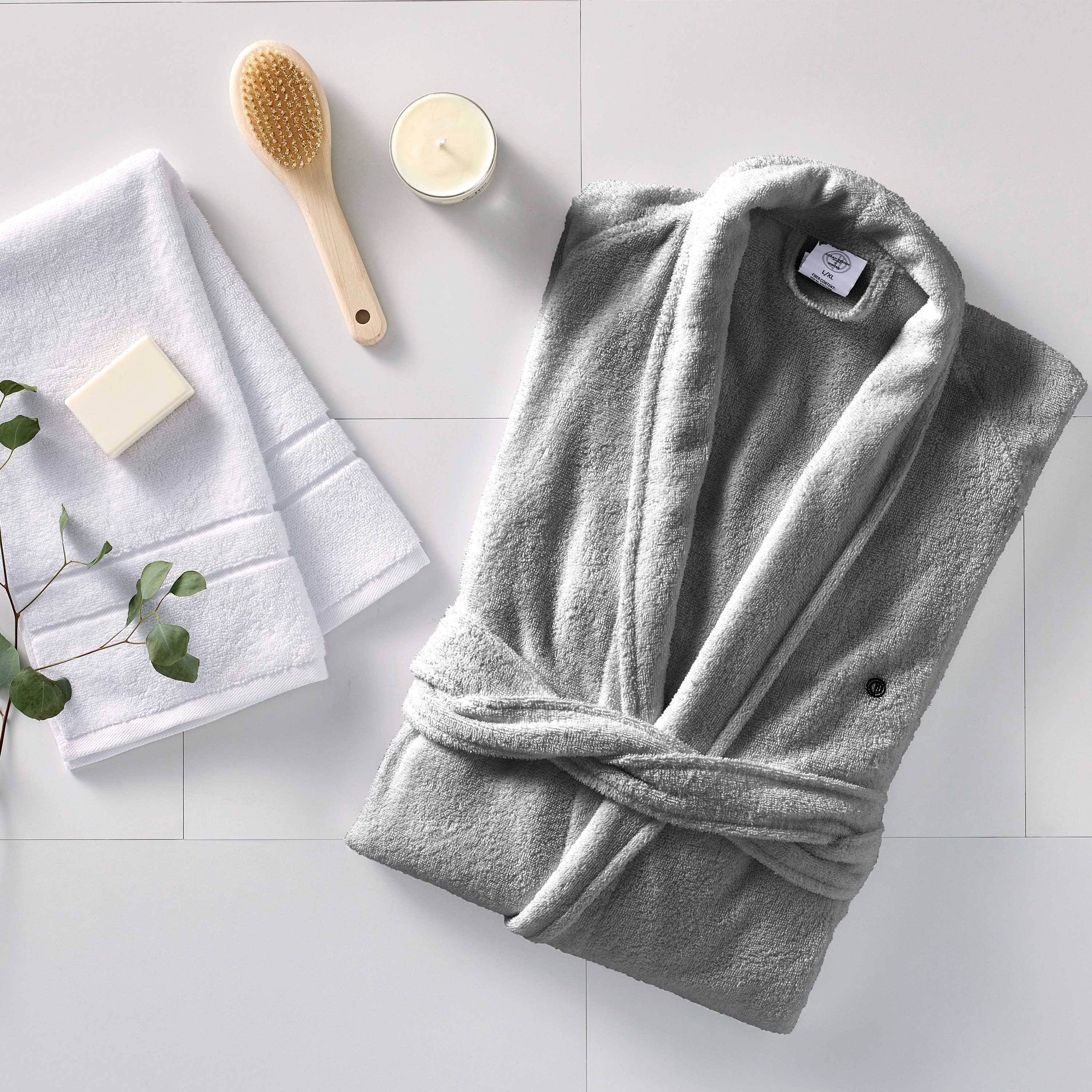Gray terry bath robe folded with white towels, a bar of soap, a wooden brush, and a candle on a light surface.