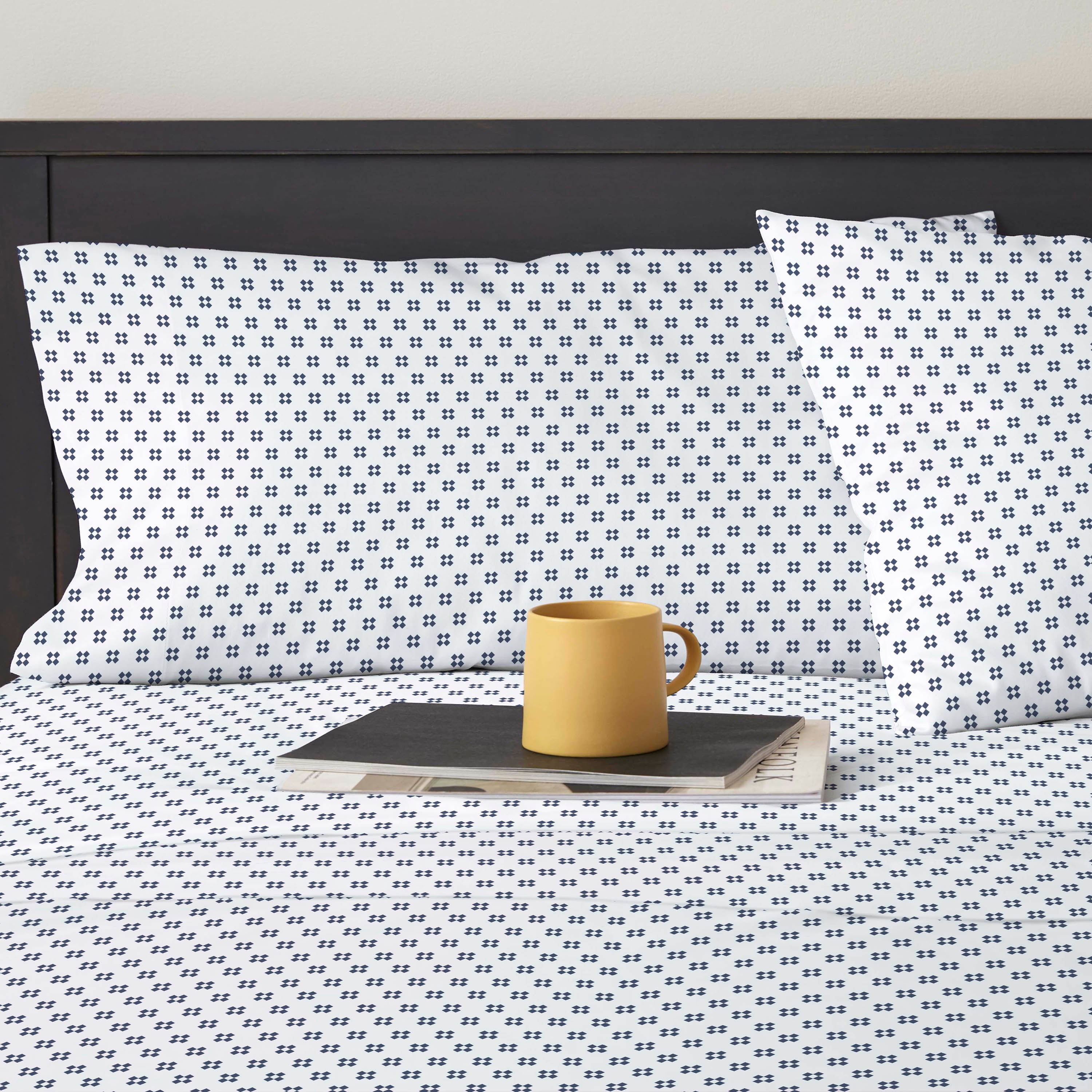 White percale sheet set with a navy blue geometric pattern, featuring two pillows and a yellow mug on a dark surface.