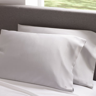 Two satin standard pillowcases in light gray positioned on a bed with a gray headboard.