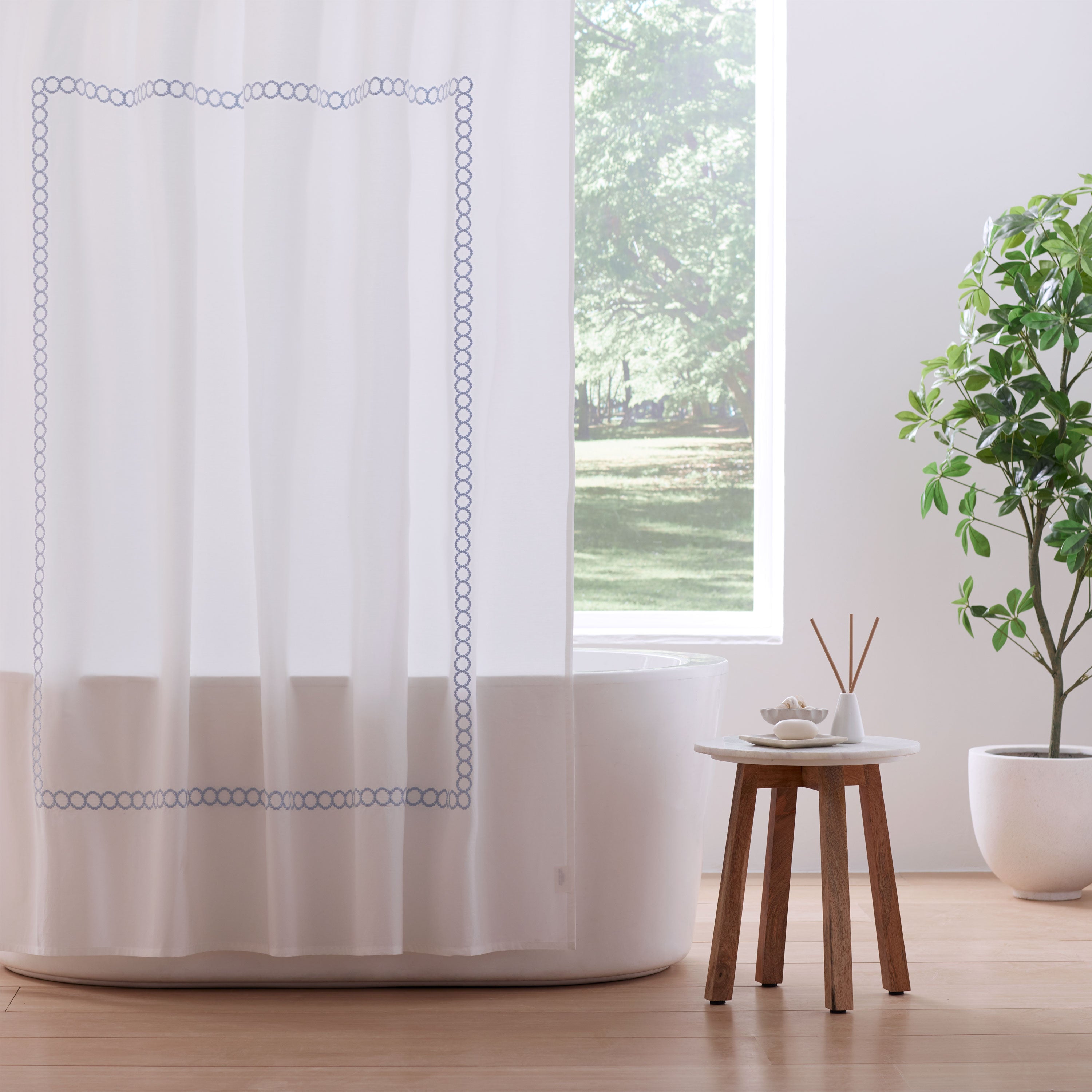 White shower curtain with blue circular border, bathtub, small table, and potted plant in a bright bathroom setting.