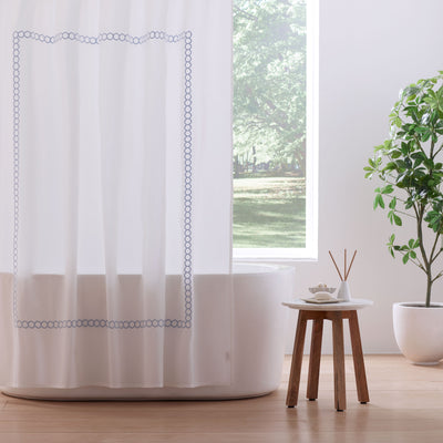 White shower curtain with blue circular border, bathtub, small table, and potted plant in a bright bathroom setting.