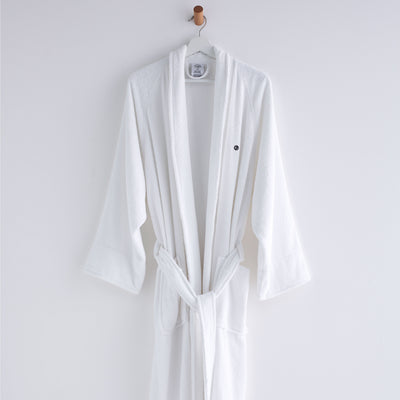 White terry bath robe with long sleeves and a tied waist, hanging on a hook against a plain background.