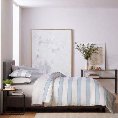 Duvet set featuring light blue and white striped design on a bed with decorative pillows and a side table.