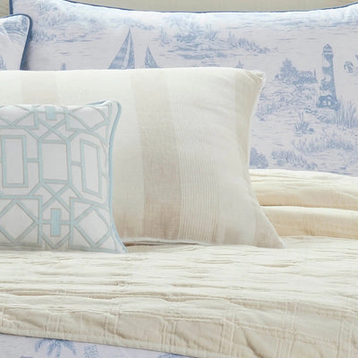 A close-up of a bed styled with bedding, featuring a cream quilted coverlet, a cream pillow with vertical stripes, a patterned cream and light blue accent pillow, and a blue toile print pillow in the background.
