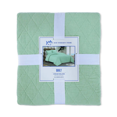 A folded sage green quilt wrapped in a white ribbon with a central paper label that reads Southern Tide Quilt Cedar Island.