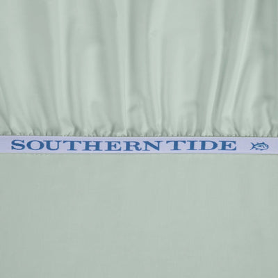 A close-up of a light sage green fabric product, likely a bed sheet or fitted sheet, gathered at the top edge with a white elastic band featuring the blue text 'SOUTHERN TIDE' and a small fish logo.