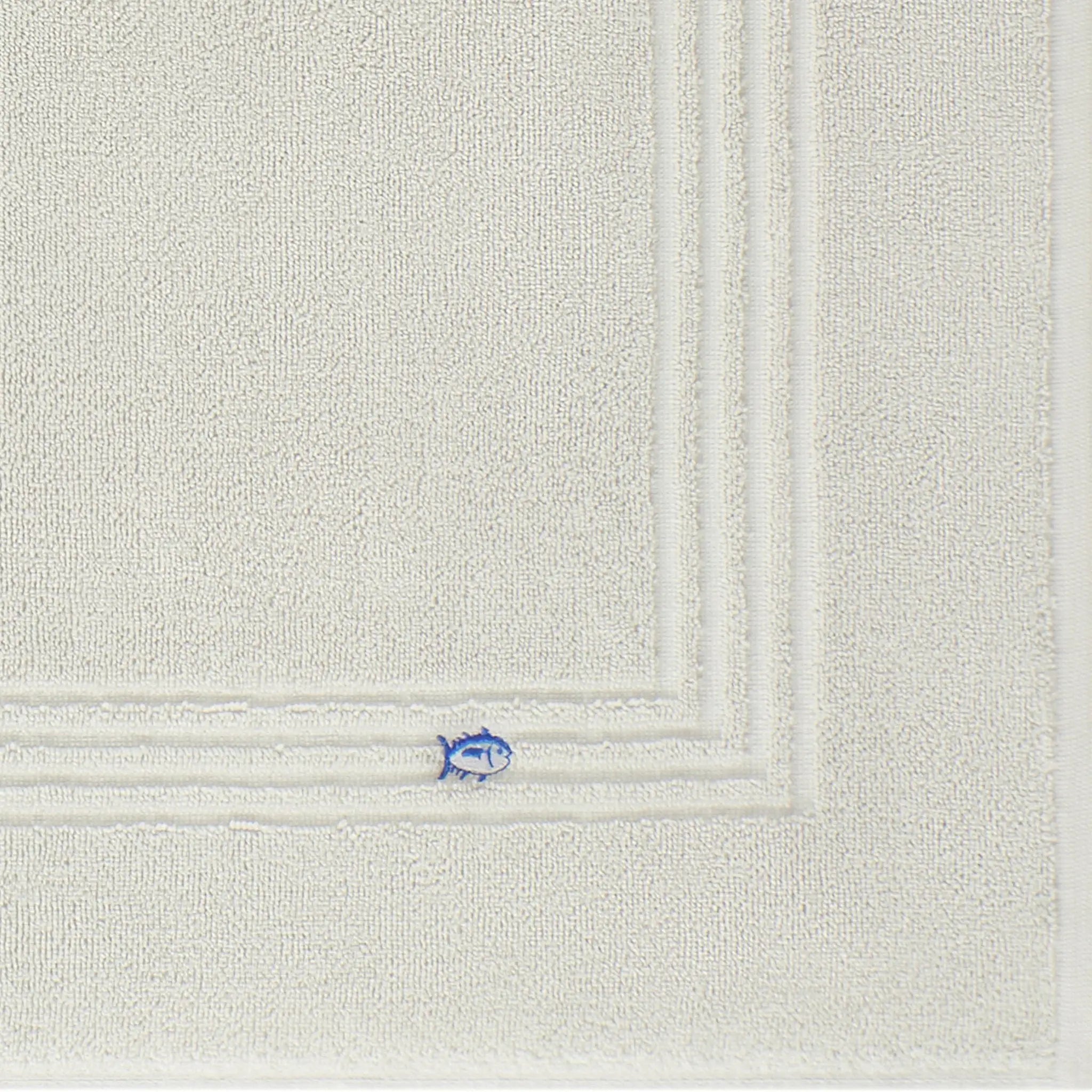 A close-up of a grey textured bath mat fabric, showing a corner with a small embroidered blue fish detail.