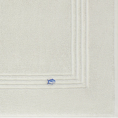 A close-up of a grey textured bath mat fabric, showing a corner with a small embroidered blue fish detail.