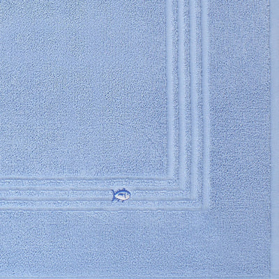 A close-up of a blue textured bath mat fabric with a stitched border and a small embroidered fish motif near the corner.