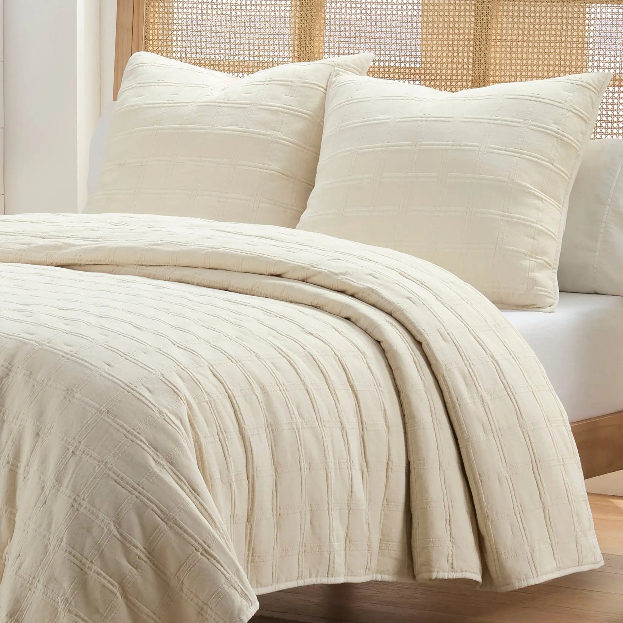 Ivory textured bedding including a quilt and two matching euros on a bed with a woven headboard.