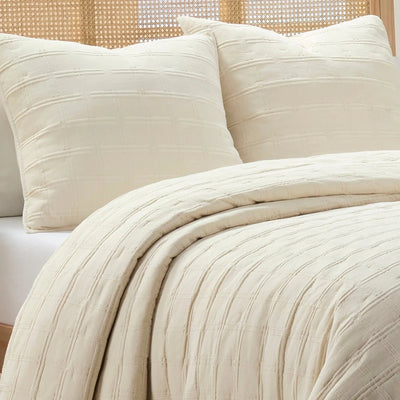 Ivory textured quilt and two matching euros with a ribbed, quilted pattern, styled on a bed with a woven cane headboard.