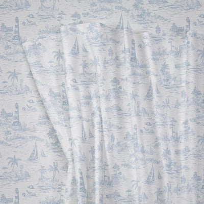 A close-up of a fabric with a light blue toile de Jouy pattern on a white background, showing draped folds and a nautical scene with sailboats, palm trees, and lighthouses.