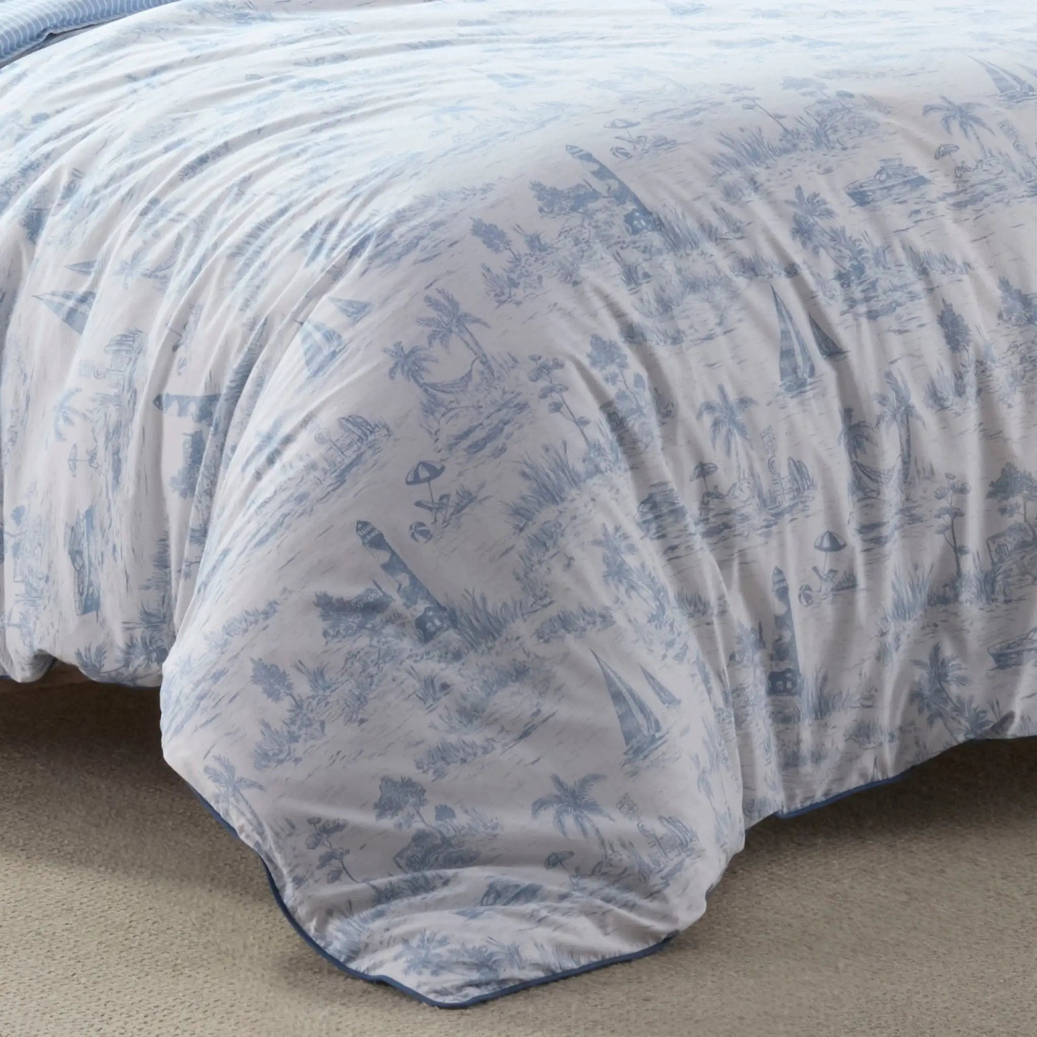 A close-up of a white comforter with a blue toile de Jouy pattern, draped over the corner of a bed on a beige carpet.