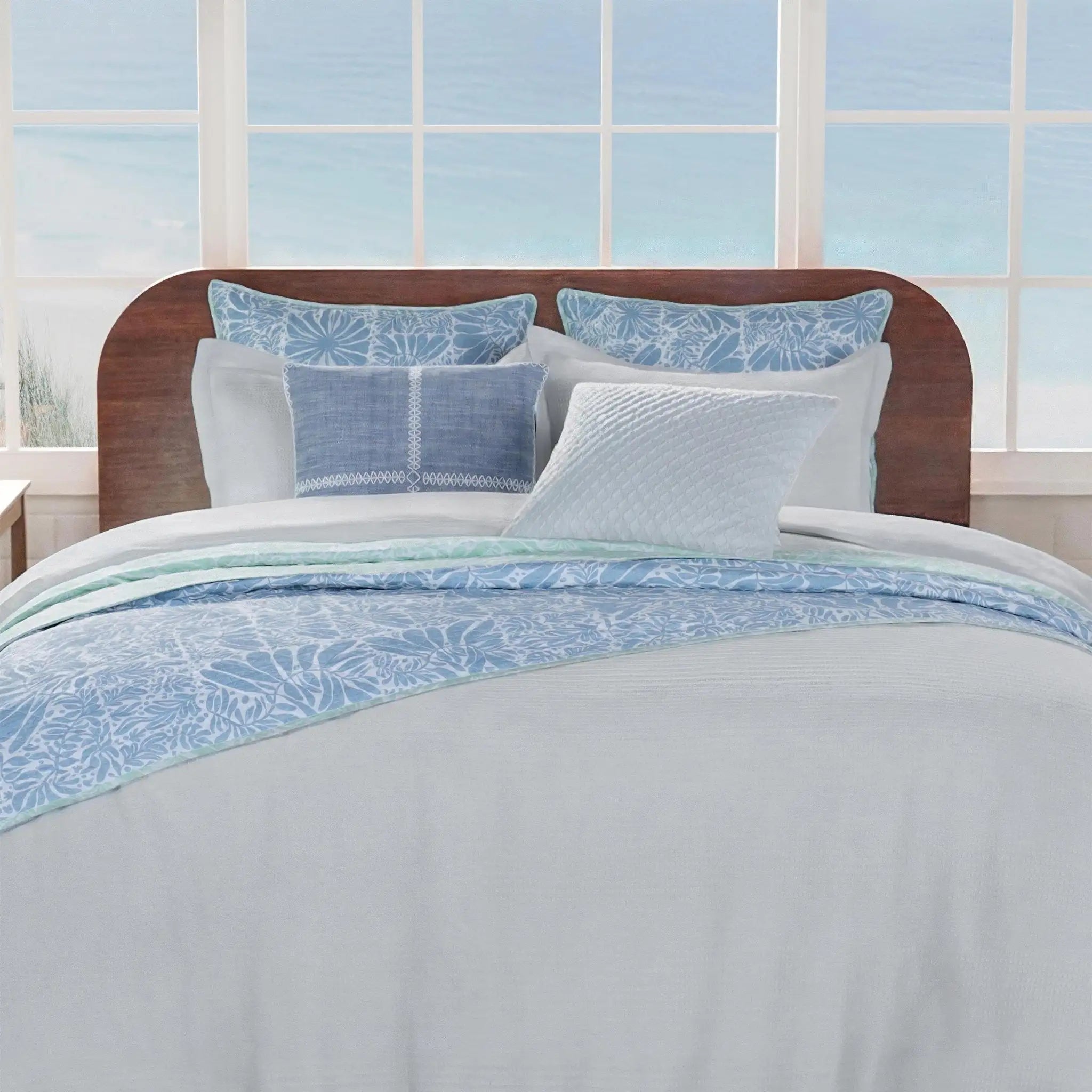 A neatly made bed with white bedding and light blue patterned pillows and throw, set in front of a large window with a beach view.