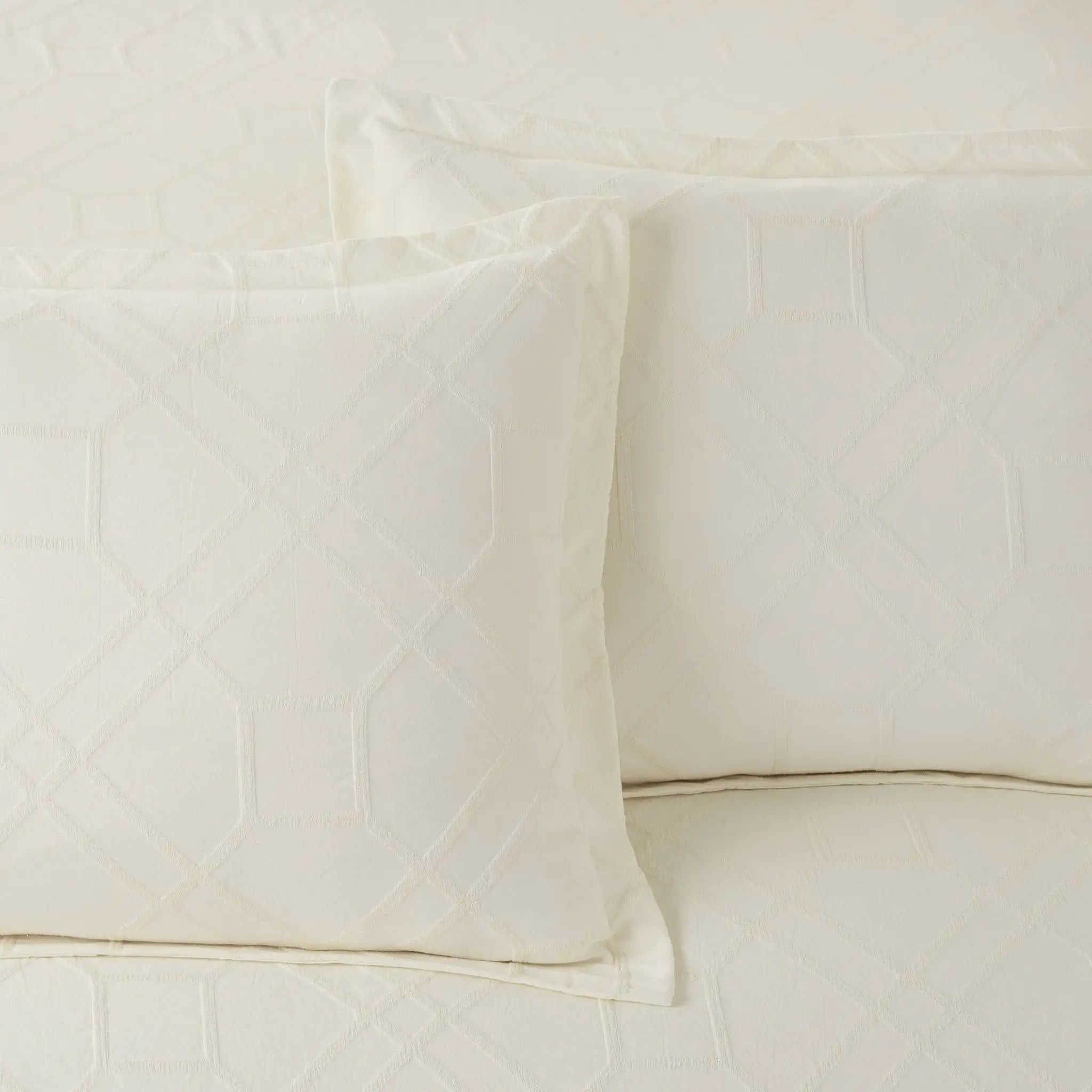 Two ivory quilted pillow shams with a geometric diamond pattern, displayed on a matching quilted bedspread.