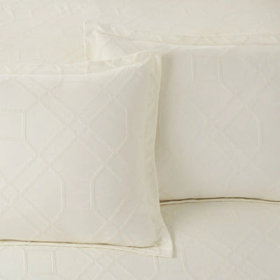 Two ivory quilted pillow shams with a geometric diamond pattern, displayed on a matching quilted bedspread.