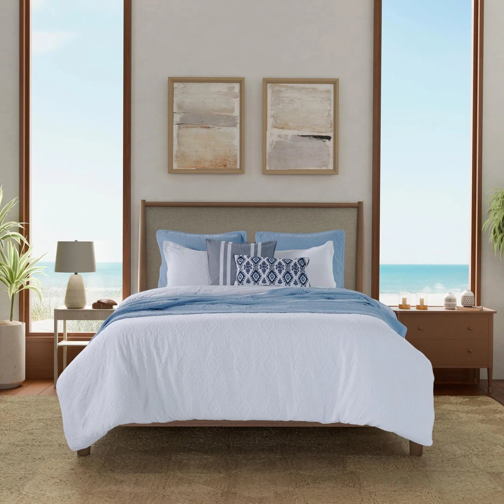 A neatly made bed with white bedding and blue pillows and throw, centered in a bright beach-view bedroom.