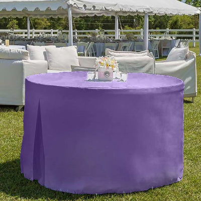 A round outdoor table covered with a violet color tablecloth, set on green grass in front of white patio furniture and a white event tent.