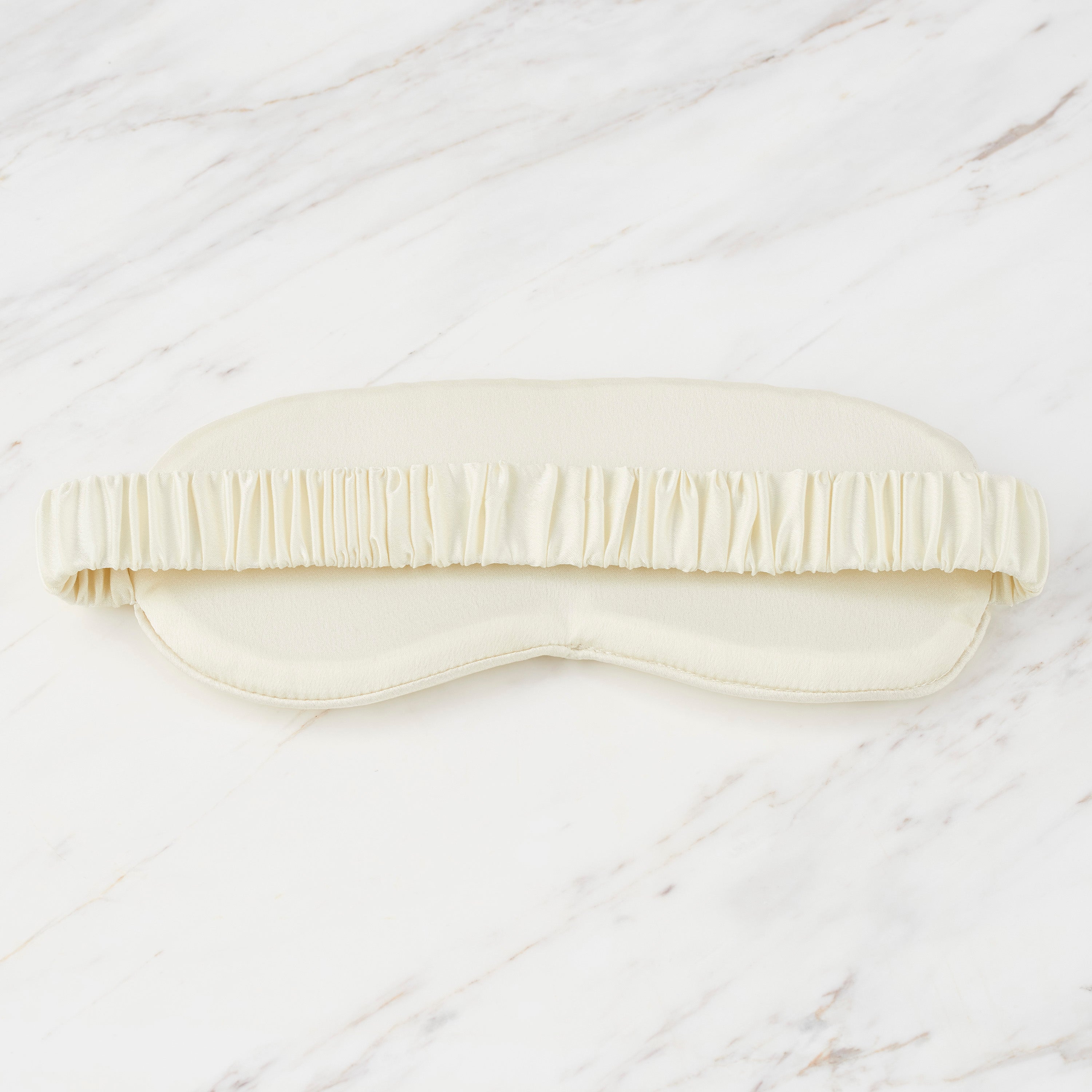 Satin eyemask with a ruffled elastic band, displayed on a marble surface.