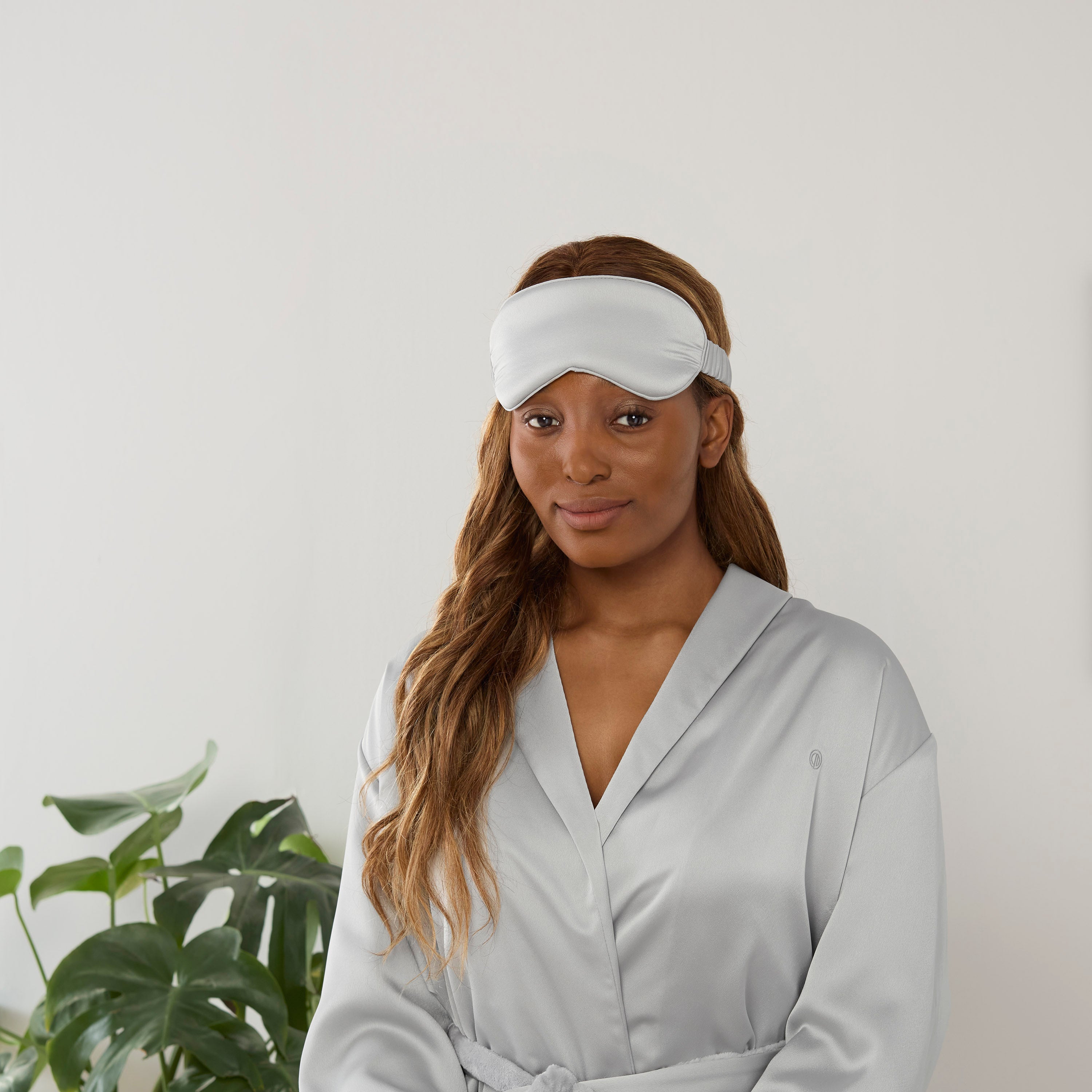Model wearing a light gray satin robe and matching eye mask, with a plant in the background.