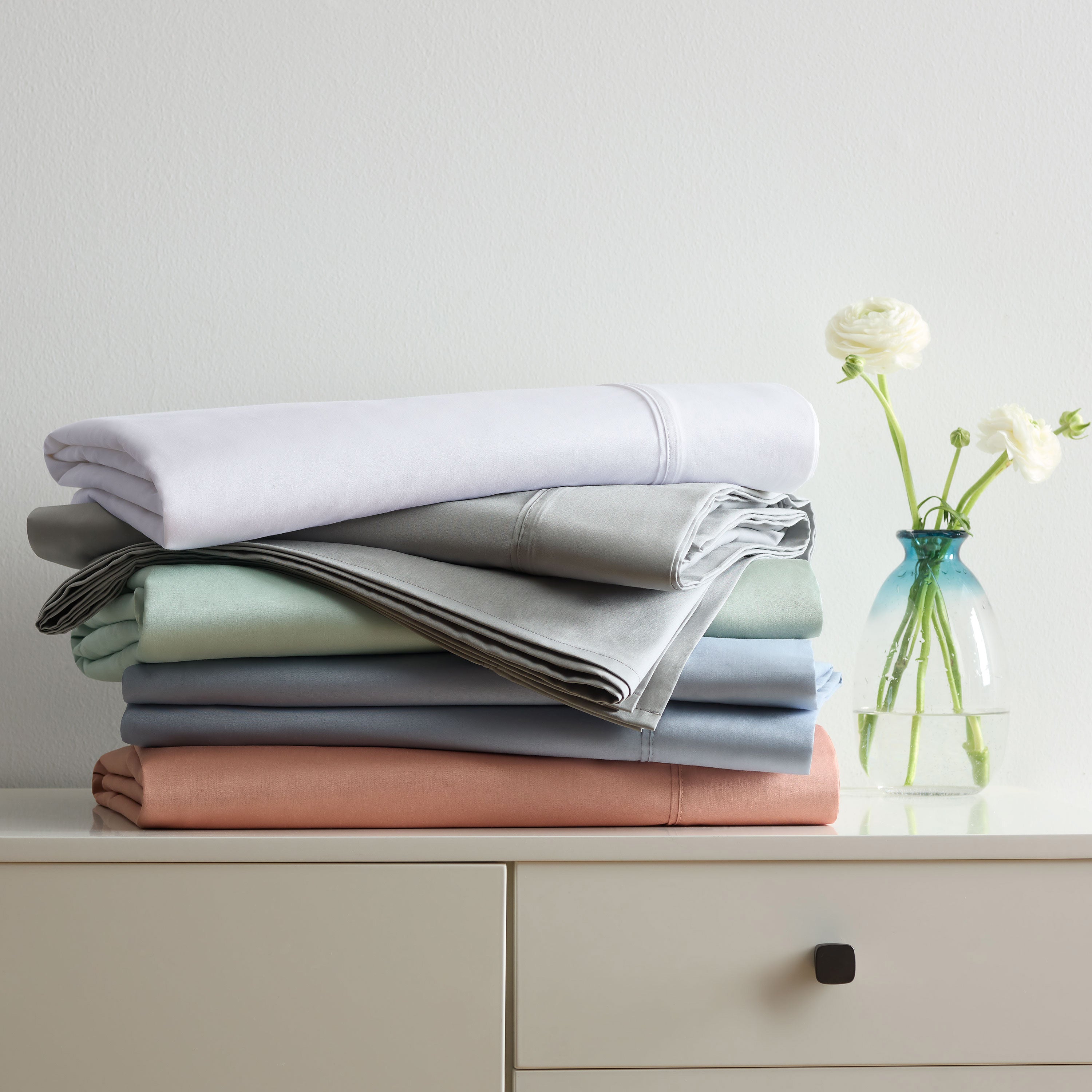Stack of solid cotton and Tencel sheet sets in various colors next to a vase with flowers on a light-colored surface.
