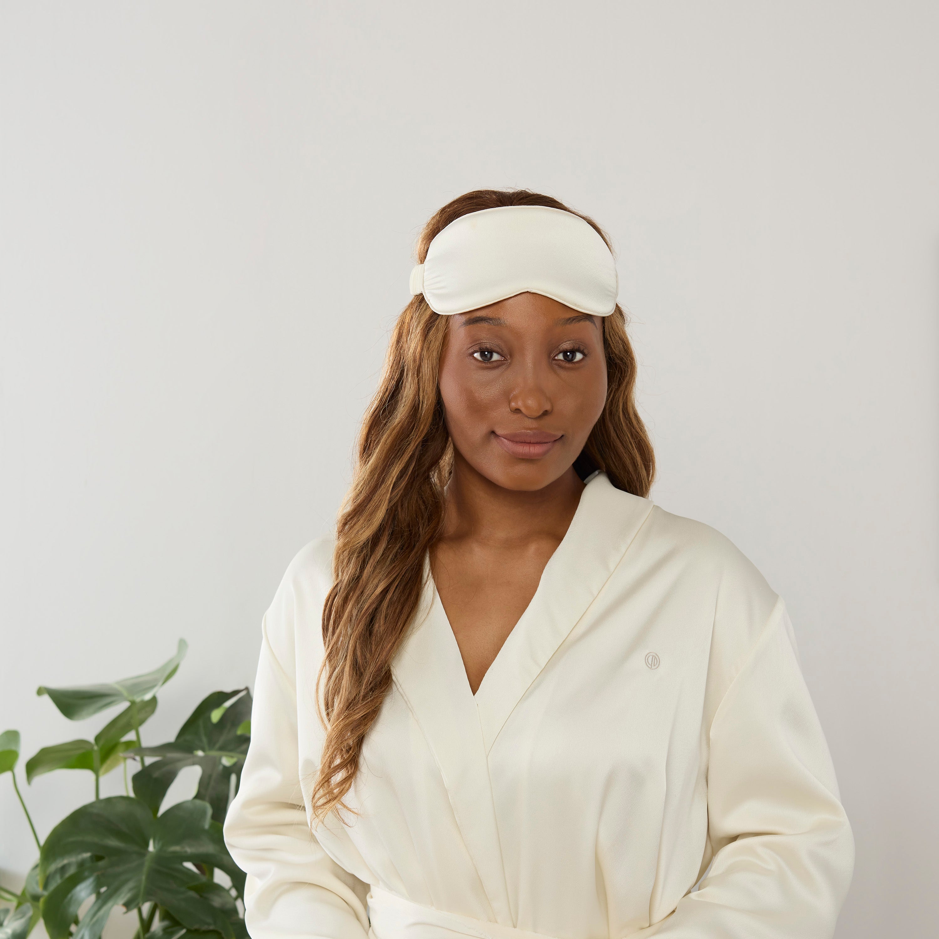 Model wearing a cream satin robe and matching eyemask, standing in front of a light background with a plant.