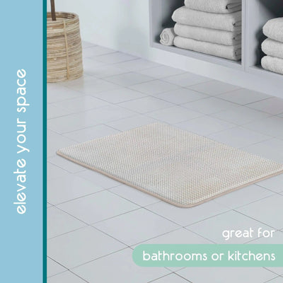 A rectangular textured sand bath rug, shown on a white tiled floor in a bathroom setting with folded towels on shelves and promotional text overlays.